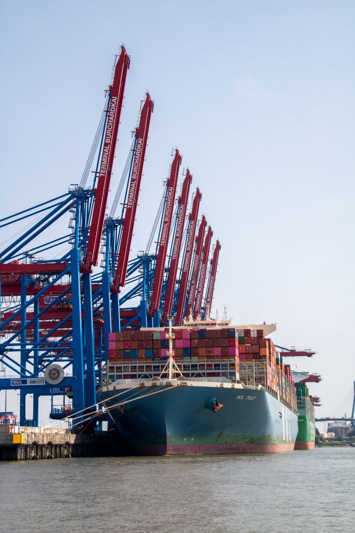 A large container ship docked at Hamburg port surrounded by harbor cranes.