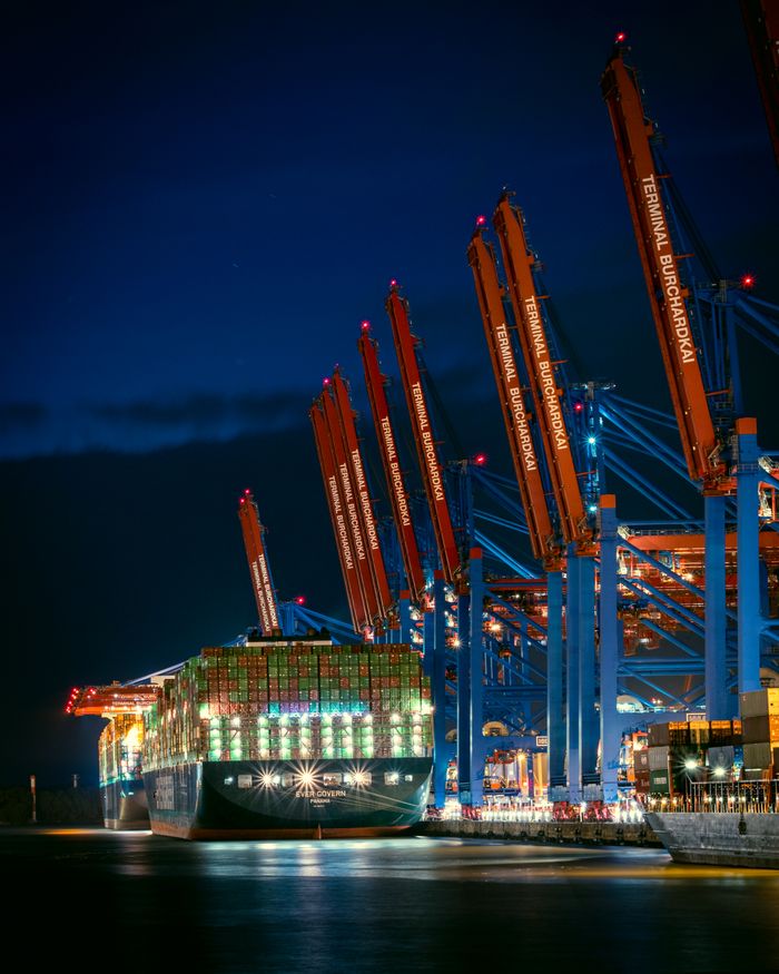 Home A large container ship docked at Hamburg's port, illuminated against the night sky.